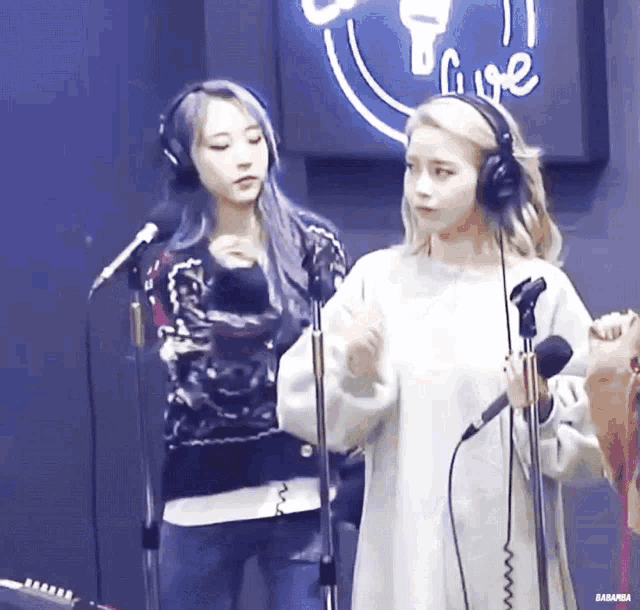 two girls wearing headphones are standing in front of microphones in a recording studio .