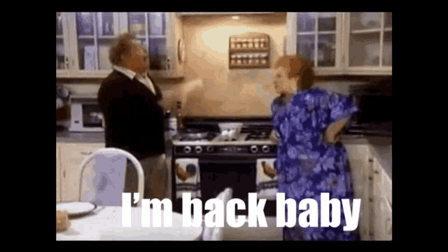 a man and a woman are standing in a kitchen with the words `` i 'm back baby '' written on the screen .