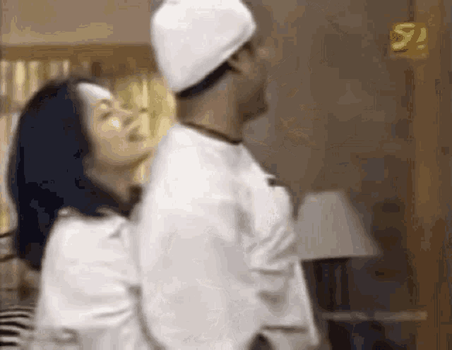 a man and a woman are standing next to each other in a room . the man is wearing a white hat .