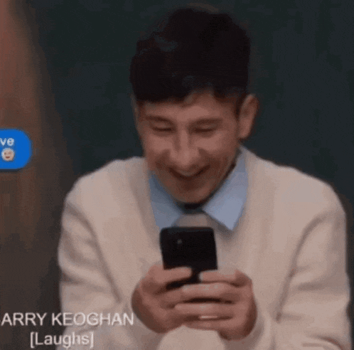 a man in a white sweater is laughing while looking at his cell phone .