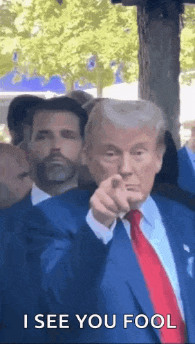 donald trump is pointing at the camera while wearing a blue suit and tie .