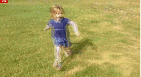 a little girl in a blue dress is running in a grassy field with a live icon above her