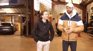 two men standing in a garage with a car parked in the background