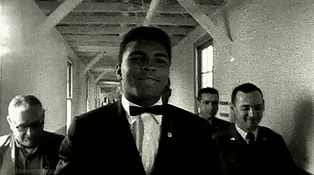 a man in a tuxedo and bow tie is walking down a hallway with other men .