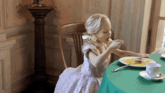 a little girl in a dress is sitting at a table with a plate of food