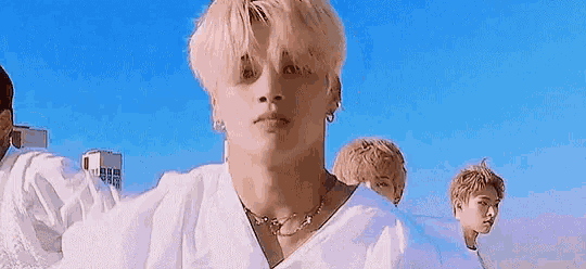 a man with blonde hair and a necklace is standing in front of a blue sky .
