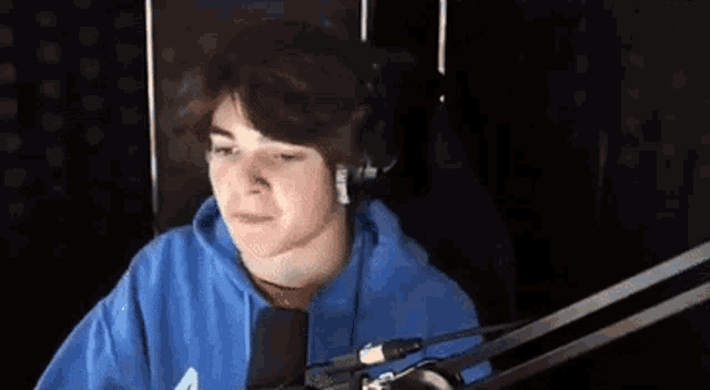 a young man in a blue hoodie is sitting in front of a microphone and wearing headphones .