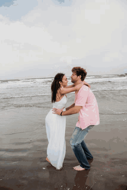 a man in a pink shirt holds a woman in a white dress on the beach