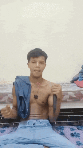 a shirtless man is sitting on a bed holding a blue shirt over his head