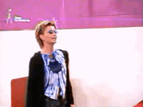 a woman wearing sunglasses and a blue and white striped shirt is standing in front of a pink wall .
