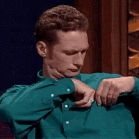 a man in a green shirt is looking at his watch while sitting in a chair .