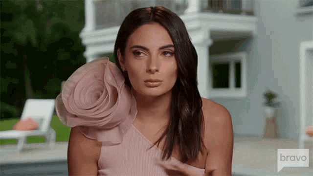 a woman in a pink dress with a flower on the shoulder is standing in front of a house on bravo .