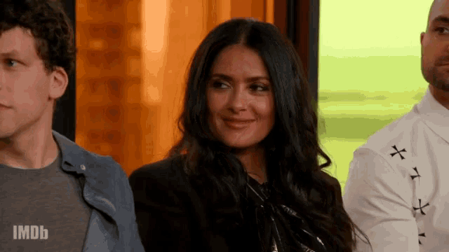 a woman in a black jacket is smiling while sitting next to a man in a white shirt with a cross on it .