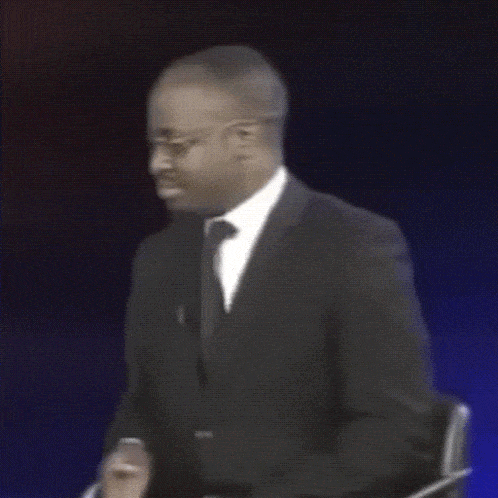 a man in a suit and tie is sitting in front of a sign that reads gay