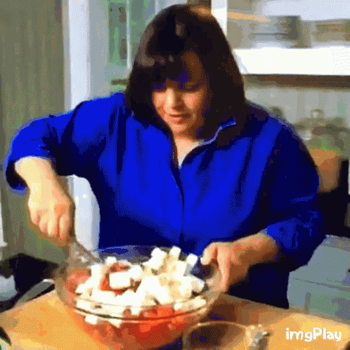 a woman in a blue shirt is mixing a salad in a glass bowl .