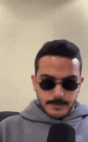 a man with a mustache wearing sunglasses and a hoodie is looking at the camera .