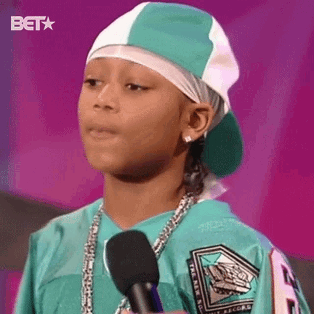 a young boy is standing in front of a microphone wearing a hat and necklace .