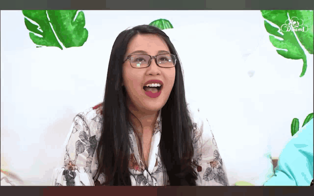 a woman wearing glasses and pink lipstick is sitting in front of a wall with green leaves on it