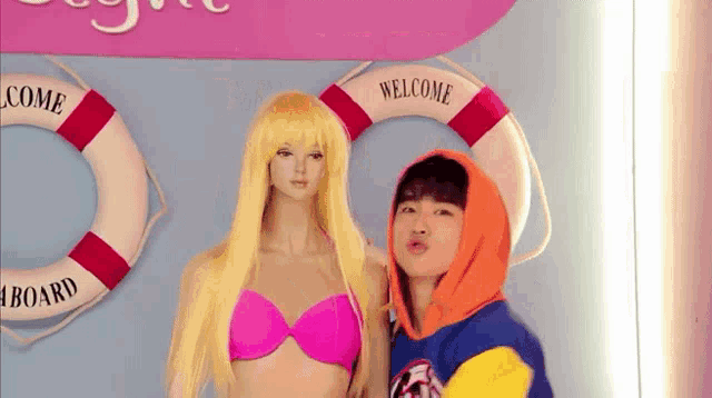 a woman in a pink bikini stands next to a mannequin with a life preserver that says welcome