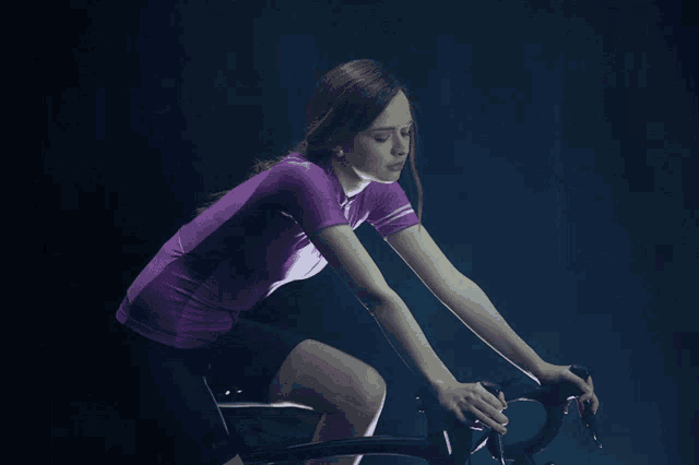 a woman in a purple shirt is riding a bike in the dark