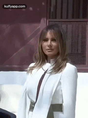 a woman in a white coat and tie is standing in front of a window and looking at the camera .