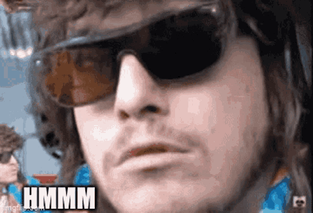 a man wearing sunglasses is making a funny face and the word hmmm is on the bottom of his face .