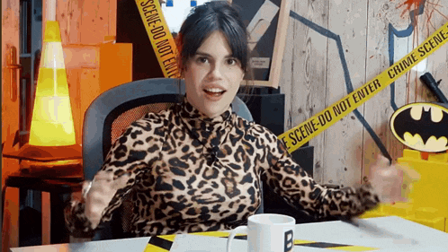 a woman in a leopard print shirt is sitting at a table in front of a batman sign and tape that says scene do not enter