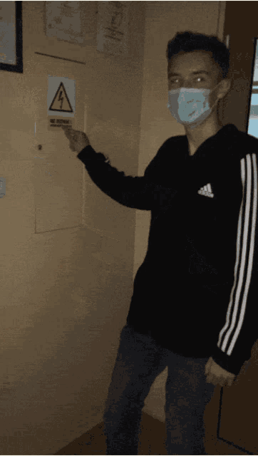 a young man wearing a mask and an adidas sweatshirt points to a sign on the wall