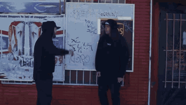 two men are standing in front of a brick building with graffiti on it including the word noro