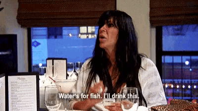 a woman sitting at a table with glasses of wine says water 's for fish i 'll drink this ..
