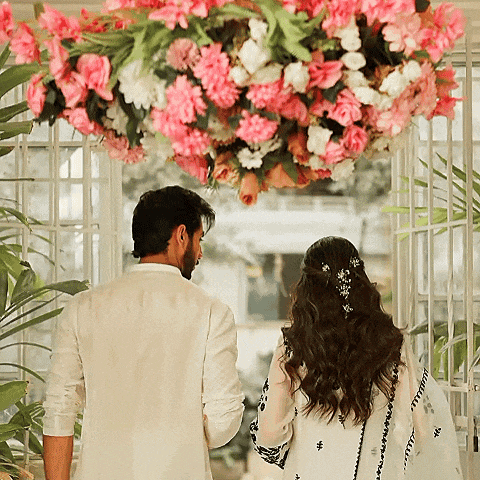 a man and a woman are standing in front of a window with pink and white flowers hanging from the ceiling