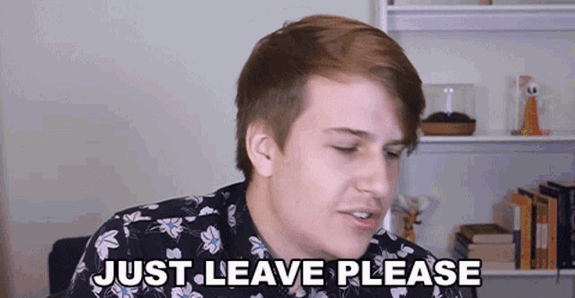 a young man in a floral shirt is sitting in front of a bookshelf and says just leave please .