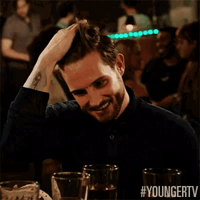 a man in a black shirt is sitting at a table with glasses of beer and scratching his head .