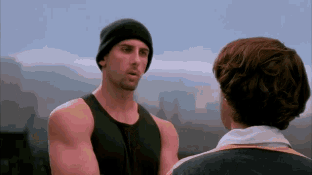 a man wearing a black tank top and a black beanie is talking to another man