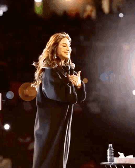 a woman in a black coat stands on a stage with a microphone in her hand
