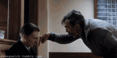 two men are standing next to each other in a room . one of the men is touching the face of the other man .