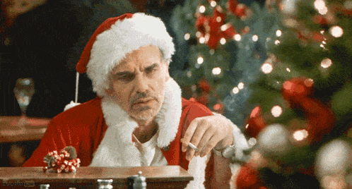 a man in a santa suit is smoking a cigarette in front of a christmas tree