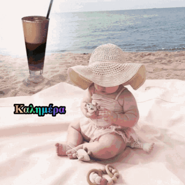 a baby wearing a straw hat is sitting on a blanket on the beach with a glass of coffee in the background