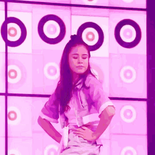 a woman in a purple shirt is standing in front of a pink wall with black circles on it