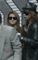 a man in a suit stands next to a woman wearing sunglasses and a hat