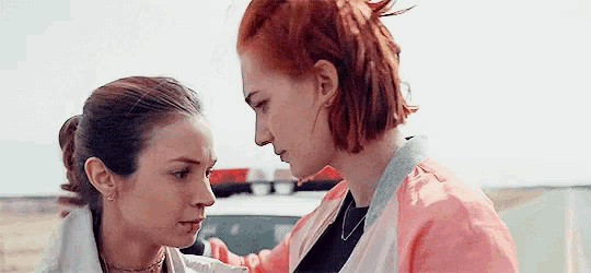 two women are standing next to each other in front of a police car and looking at each other .