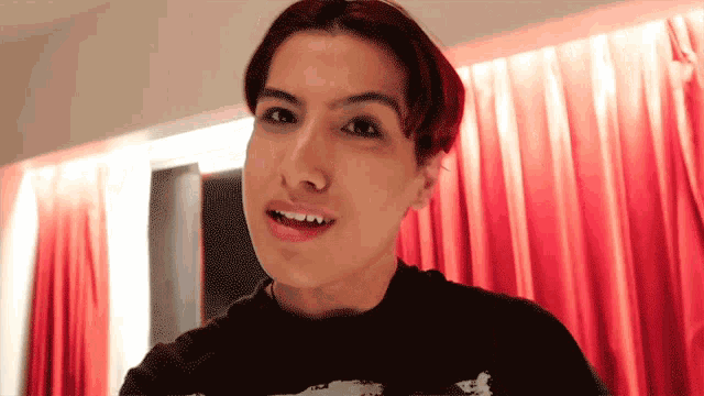 a young man with red hair is smiling in front of a red curtain behind him