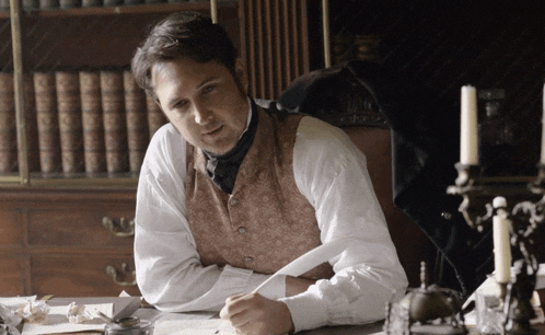 a man in a vest sits at a desk with candles in the background