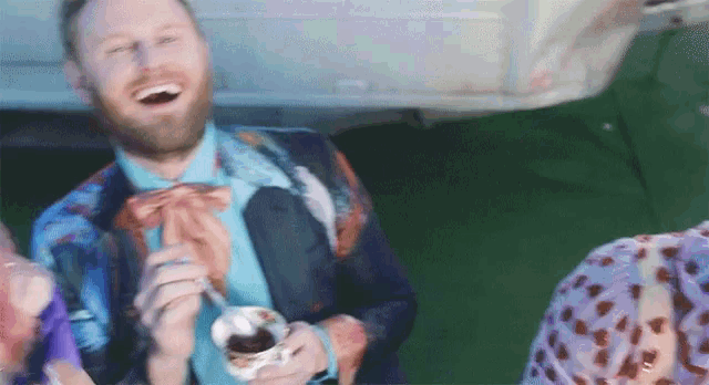 a man in a suit and bow tie is laughing and holding a cup of ice cream