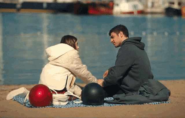 a man and a woman sit on a blanket on the beach looking at each other