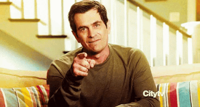 a man is sitting on a couch pointing at the camera with a city tv logo on the couch