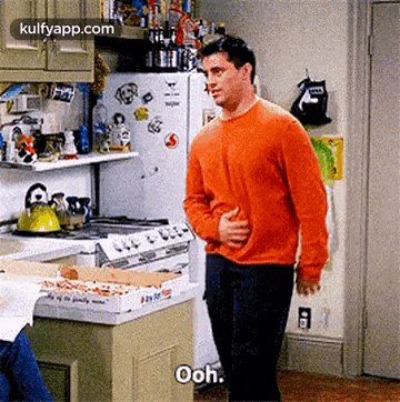 a man in an orange sweater is standing in a kitchen next to a pizza box and a refrigerator .