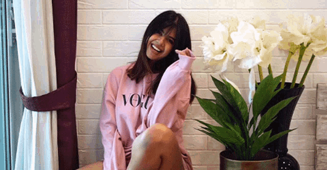 a woman wearing a pink sweater that says vogue sits next to a potted plant