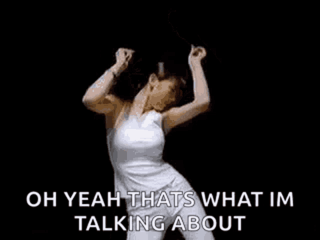 a woman is dancing in a white tank top and white pants in a black room .