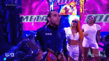 a man in a blue jacket with the word dar on it is standing next to two women in white dresses .